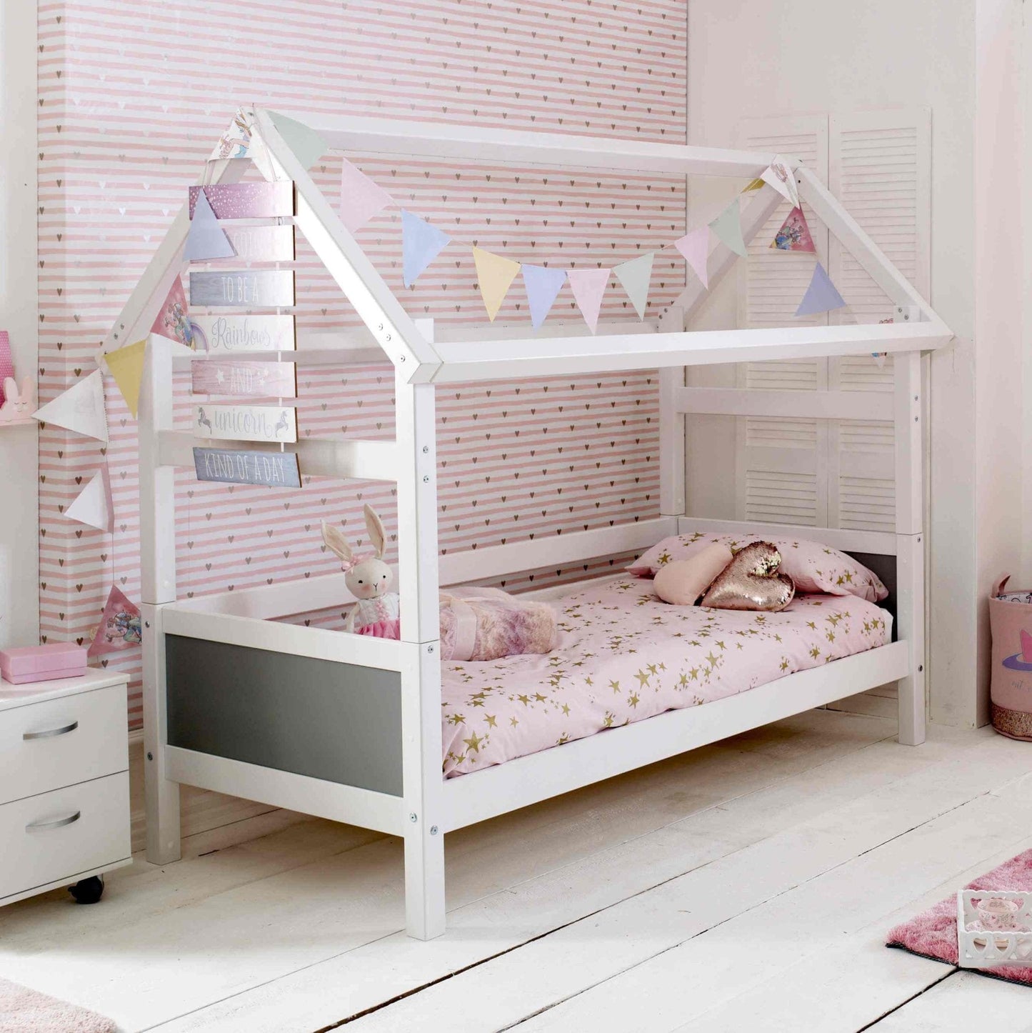 Kids Nordic Playhouse Bed with optional trundle and storage drawers, featuring a house frame, colorful bunting, and a cozy pink bedding set.