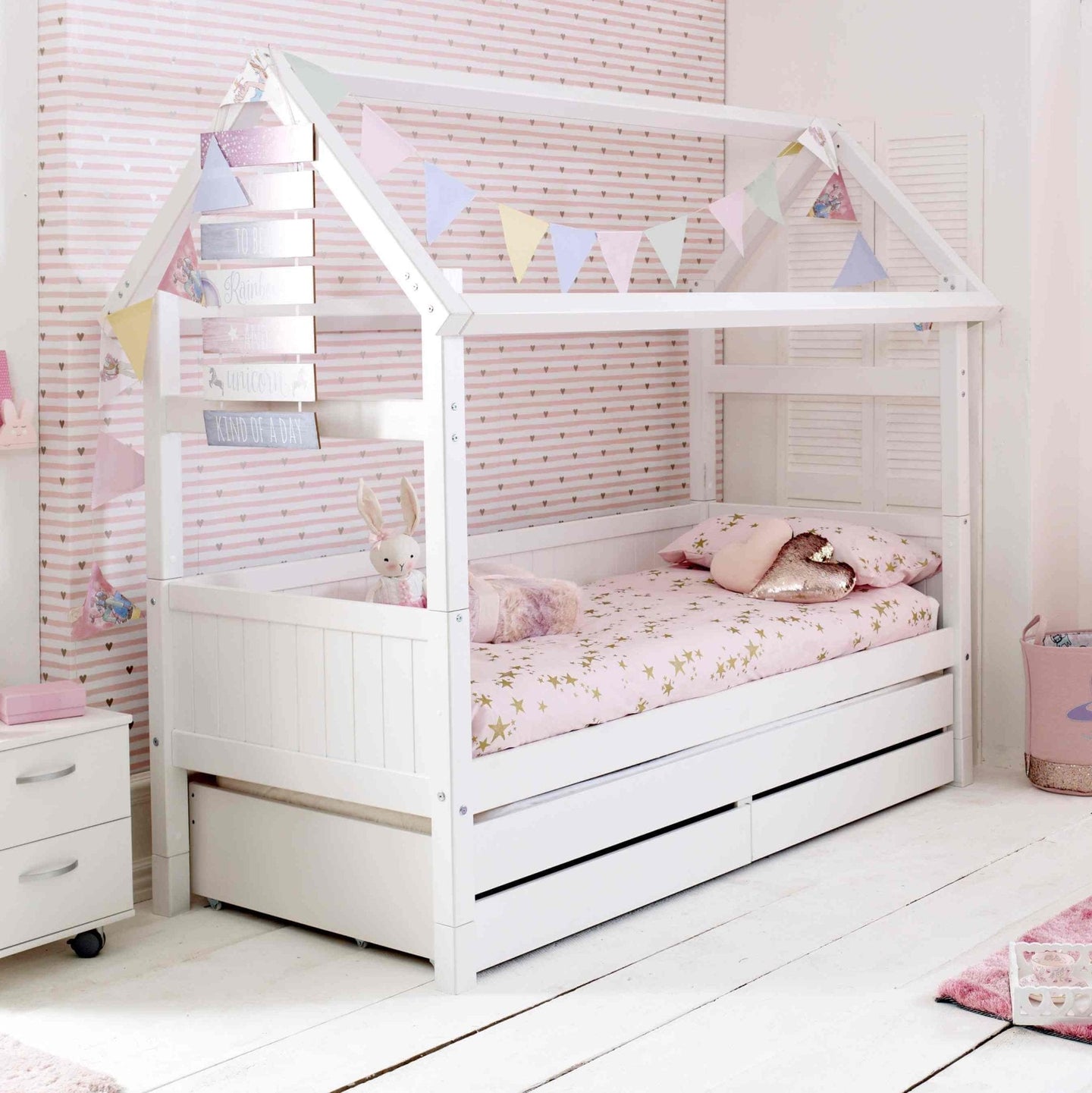 Nordic playhouse bed with optional trundle and storage drawers in children's room, white finish, decorated with colorful bunting.